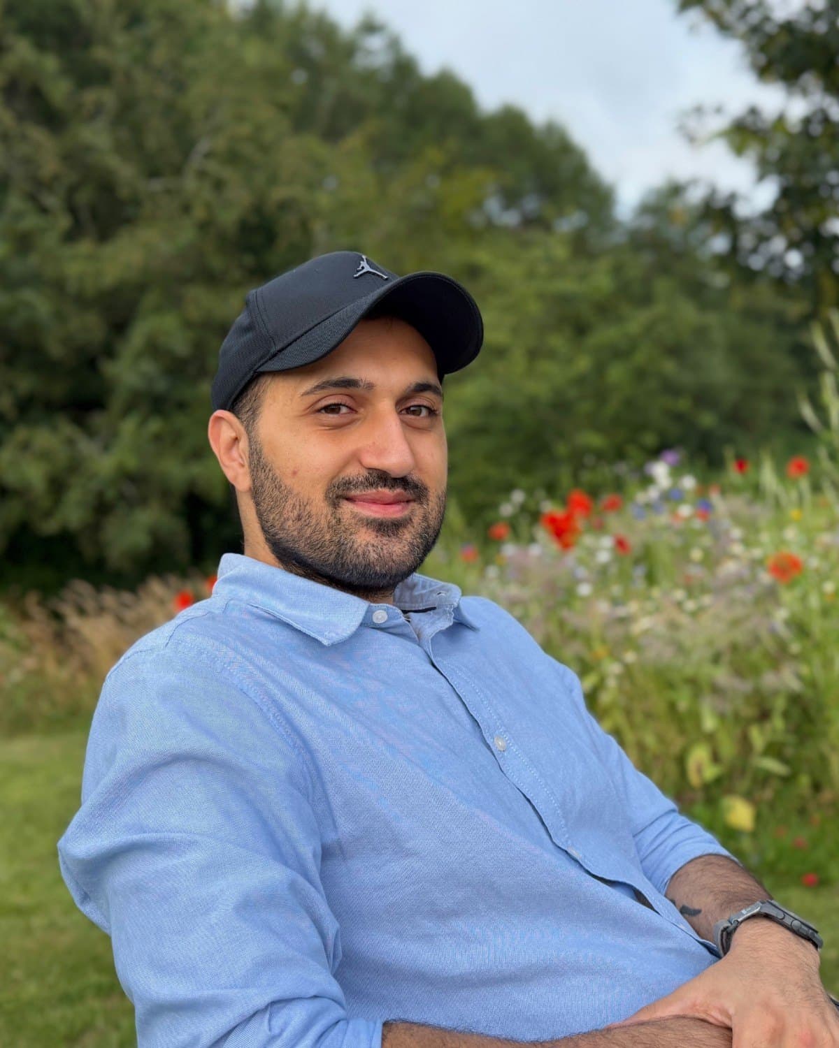Mo Ghasemi in a blue shirt and cap, sitting in a garden of wildflowers, smiling at the camera