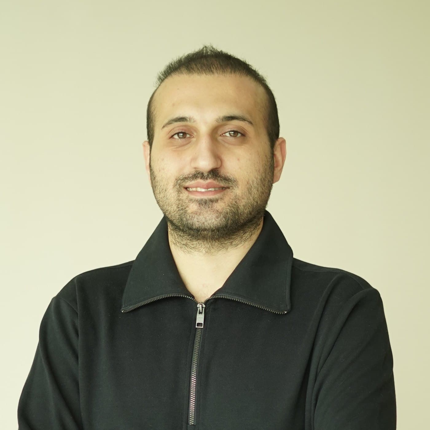 Mo Ghasemi, smiling, in a black zip-neck top against a plain cream wall